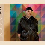 A man in a black vest over a black shirt in a black hat standing in front of a multi-colored abstract painting in the works.