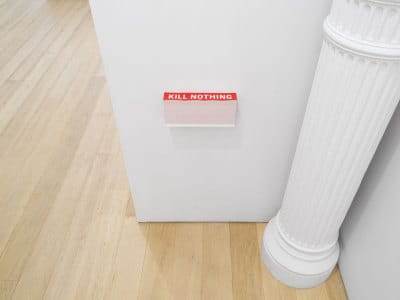 A stack of stickers that read 'KILL NOTHING' in white block letters on a red background. They rest on a wood shelf floating in the middle of a wall.