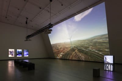 View of a museum exhibition with a large projection at the center and smaller screens on either side. 