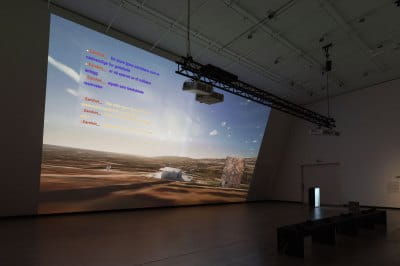 View of a large screen in a museum gallery showing the landscape of Golan Heights with chat text from a video game overlaid. 