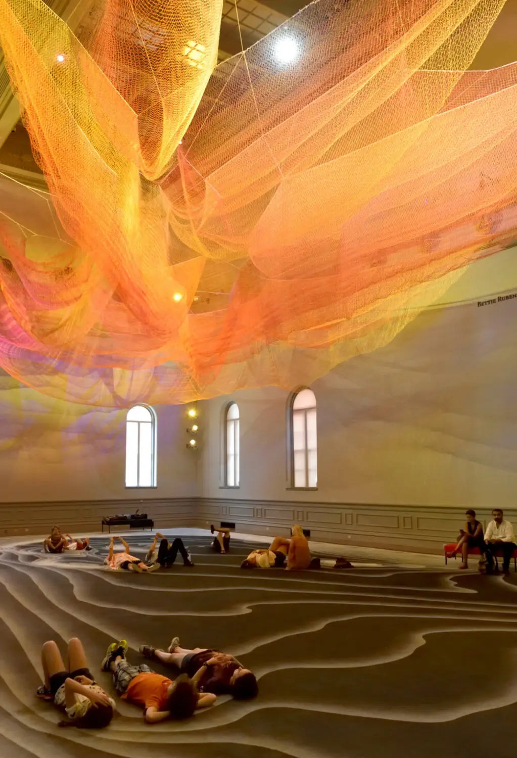 people lay underneath a green and pink Janet Echelman installation suspended from the ceiling