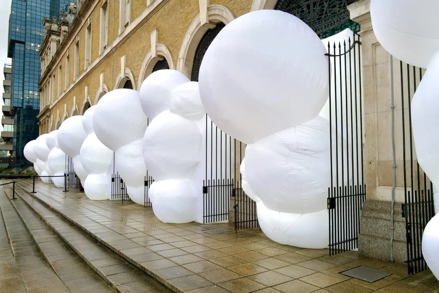 a side view of a series of large, white, bulbous, inflatable shapes designed by Steve Messam that stick out of an arched arcade on the first level of a brick building